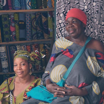Two women sitting next to a shelf filled with colorful fabric